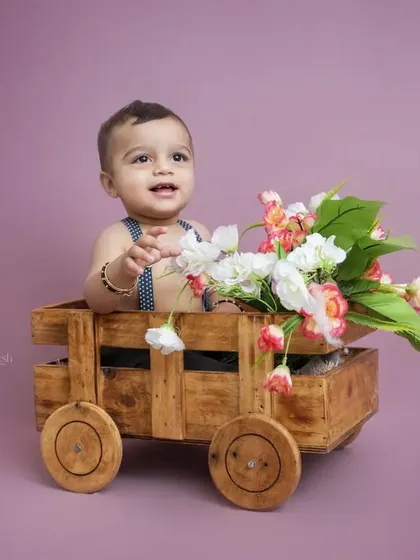 A sweet baby boy sits in a wooden cart filled with flowers. The soft purple background complements the floral arrangement and creates a gentle, lovely portrait.