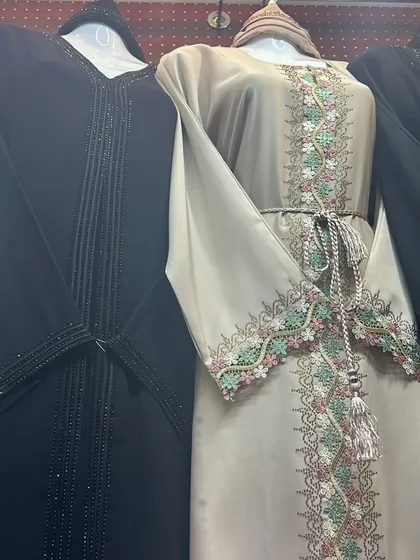 On the left, a classic black abaya with simple vertical beading. On the right, a beige abaya with colourful floral embroidery and a matching tassel belt.