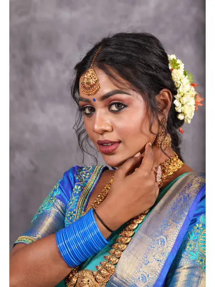 A close-up portrait focusing on the vibrant colors of a blue and teal silk saree. The shot highlights the model's makeup, gold jewelry, and the bright blue bangles, creating a cohesive and striking look.