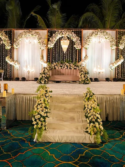 A wide view of an elegant reception stage. The backdrop of white drapes and fairy lights, combined with floral arches and chandeliers, creates a sophisticated and romantic setting.