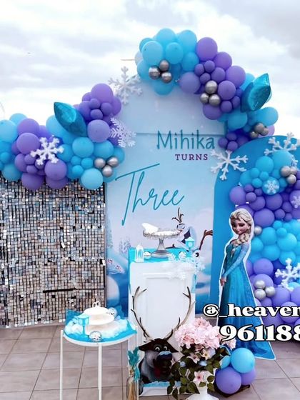 A grand Frozen-themed setup on a rooftop for a third birthday. The decor features a silver sequin backdrop, character cutouts of Elsa and Olaf, and a balloon arch in shades of blue, purple, and silver.