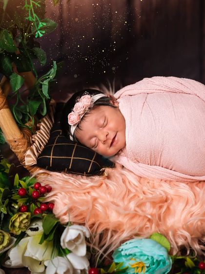 A smiling newborn sleeps on a soft pink fur blanket, surrounded by flowers. The added sparkle effect gives this photo a magical touch.