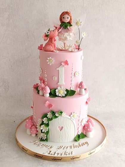 A sweet fairy-themed cake for Advika's first birthday. The two-tiered pink cake features a fairy house, mushrooms, and a cute little dragon.