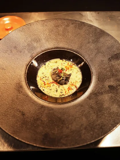 A modern Indian dish served in a dark, textured bowl, where a central element is surrounded by a light, creamy curry.