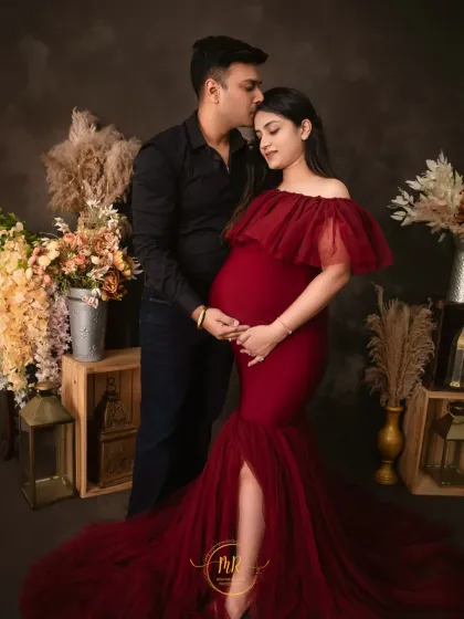 A tender moment between partners. The expecting mother, dressed in a stunning off-the-shoulder red gown, shares a loving glance with her partner, who kisses her forehead in our Gurugram studio.