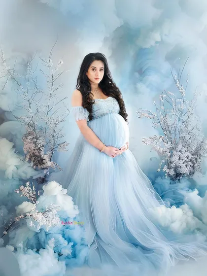 A stunning, wide-angle view of a dreamy maternity shoot. The expecting mother stands in a flowing blue gown, surrounded by a set designed to look like a beautiful sky with clouds and blossoms.