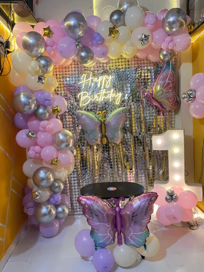 A whimsical butterfly themed first birthday party. The decor features a silver sequin backdrop, a pink and purple balloon arch, and large butterfly foil balloons.