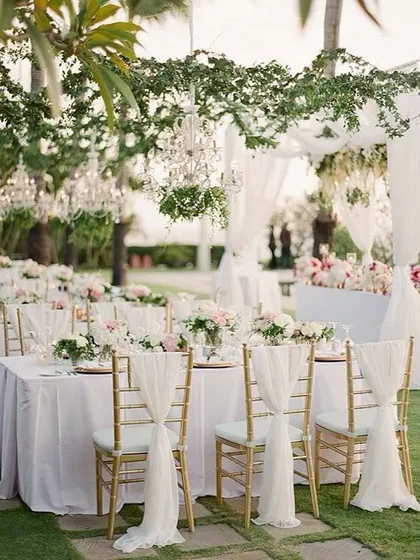 An elegant outdoor wedding reception setup. The tables are beautifully set with floral centerpieces, and the chairs are decorated with flowing white drapes.