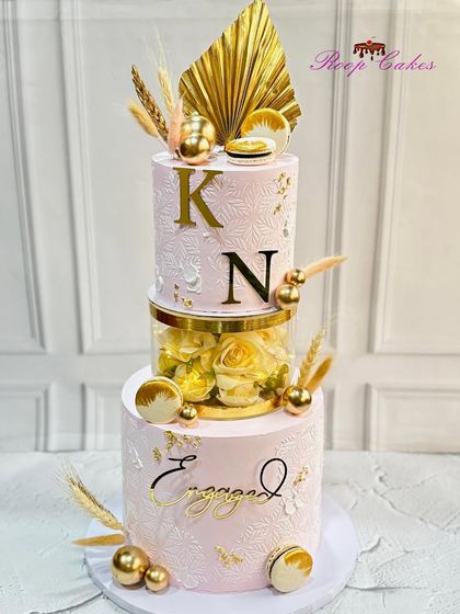 A beautiful engagement cake featuring a floating tier filled with yellow roses. The design is accented with gold palm leaves, macarons, and the couple's initials, creating a luxurious and celebratory feel.