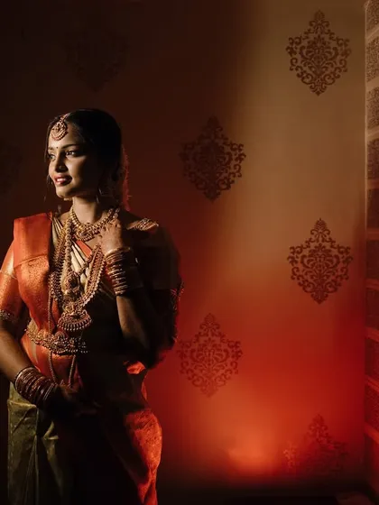 A dramatic bridal portrait using strong side lighting. The bride stands against a textured wall, her traditional South Indian attire glowing in the warm light.