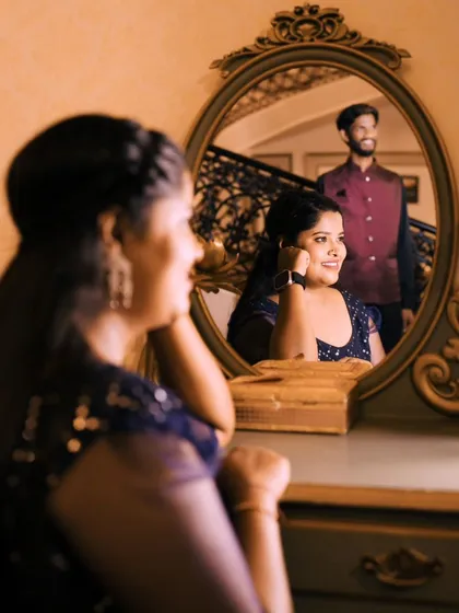A creative reflection shot. The bride looks at her reflection in a mirror, with the groom visible in the background. A very artistic pre-wedding photo.