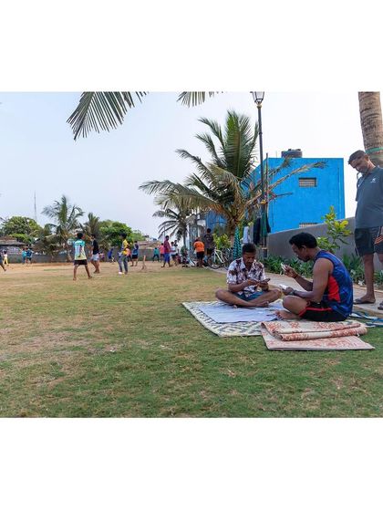 A wide view of the Gora Dabkal ground shows its multi-use nature. While a game of cricket unfolds in the background, others find a quiet spot on the grass for conversation, demonstrating the landscape's ability to support diverse activities simultaneously.