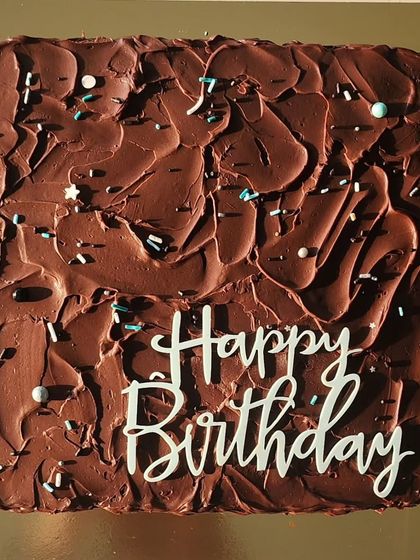 A square chocolate cake covered in a thick, rustic layer of dark chocolate ganache. The sprinkles and simple "Happy Birthday" plaque make it a perfect choice for a straightforward, delicious celebration.