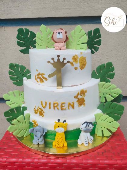 A wild jungle-themed cake for a first birthday. This two-tier cake is decorated with fondant jungle leaves and cute, hand-sculpted animals like a lion, giraffe, elephant, and zebra.