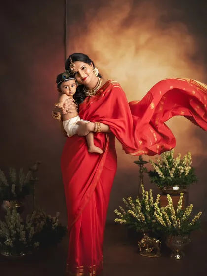 A mother and her baby Krishna, captured in a timeless, artistic portrait. The flowing red saree adds a dramatic and beautiful touch.