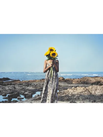 A whimsical shot for the 'Sacred Wrap' campaign for Adiv x Kavita Parmar. The styling is playful, with the model's face hidden by sunflowers, set against the raw, natural beauty of a rocky shoreline.