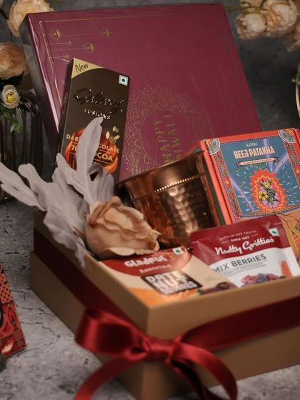A classic Diwali gift box with a rich red ribbon. This hamper contains a mix of traditional and modern treats, including a copper diya and a box of fine chocolates.