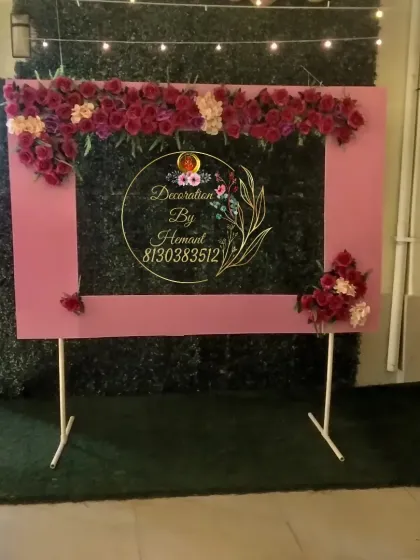 A large, custom-built selfie frame for a birthday party. The pink frame is decorated with red and pink roses and set against a green hedge wall, providing a beautiful and interactive photo opportunity for guests.