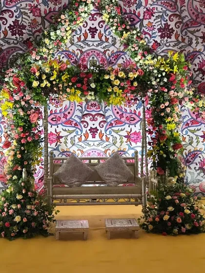 A beautiful Mehendi seating area with a floral-draped swing set against a vibrant, patterned backdrop, flanked by elegant white peacock statues.