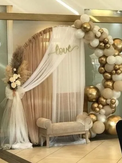 A trendy and romantic photo booth arch combining a rustic wooden frame, a cascade of neutral-toned balloons, and delicate drapery with a "love" sign, perfect for creating memorable guest photos.