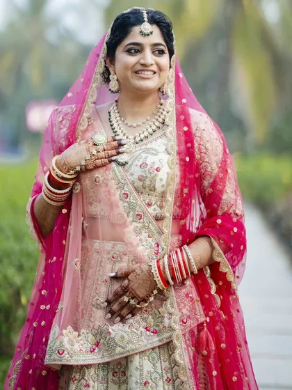 A beautiful bride ready for her sunset wedding in Goa. The makeup features a soft glam eye and a rosy lip, complementing her intricate ivory and pink lehenga. Her hair is styled in a floral updo, perfect for an outdoor ceremony.