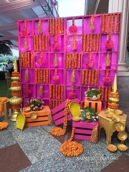 This backdrop is a symphony of tradition. A wall of pink shelves holds rows of marigold garlands and tassels, creating a textured and incredibly vibrant photo opportunity for the Haldi ceremony.
