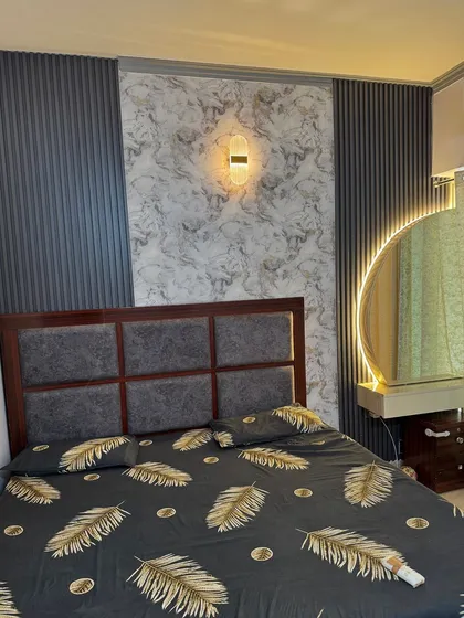 This bedroom features a dark wood bed with a grey upholstered headboard, set against a wall with marble and fluted paneling. A round backlit mirror serves as a dressing area.
