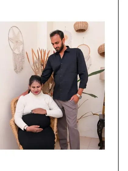 A sweet and supportive couple's portrait. The partner stands behind the seated mom-to-be, creating a sense of protection and togetherness. The natural textures of the wicker chair and wall hangings enhance the warm atmosphere.