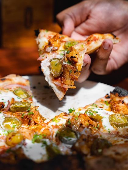 A hand holds up a slice of the BBQ Chicken Pizza, showing a cross-section of the toppings and the light, airy structure of the sourdough crust.