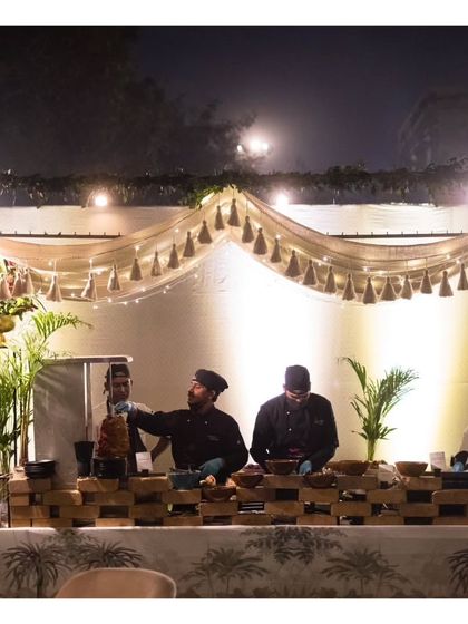 Two chefs work behind a beautifully decorated live station at an outdoor evening event. The rustic yet chic setup with tassel drapery and warm lighting creates an inviting and stylish food destination for guests.