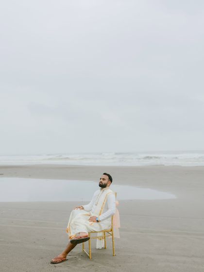 A stylish, contemplative portrait of the groom sitting on a chair on the beach. The vast, empty space around him creates a sense of peace and anticipation before the ceremony.