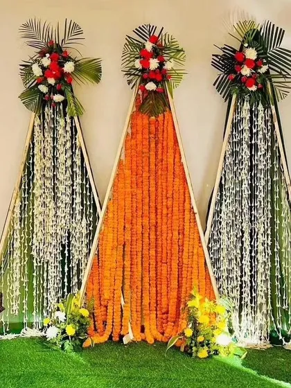 A modern Haldi backdrop with three A-frame structures. The frames are decorated with strings of marigolds and tuberoses, with floral arrangements on top, set on an artificial grass carpet.