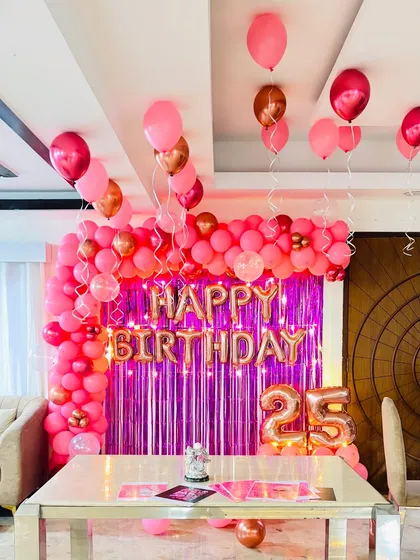 A vibrant 25th birthday setup at home. The decor includes a pink balloon arch, a pink tinsel backdrop, and rose gold marquee numbers.