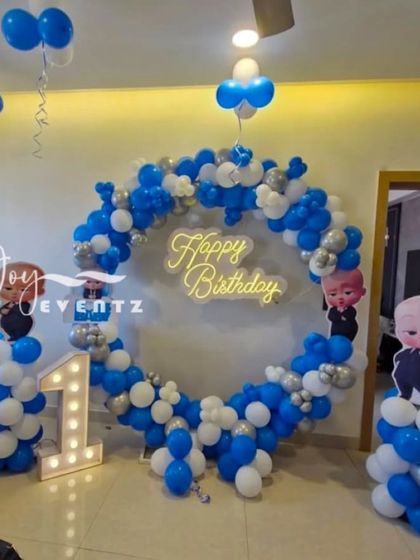 A circular balloon ring is a great option for home parties. This Boss Baby first birthday decoration uses a full ring of blue, white, and silver balloons, a 'Happy Birthday' neon sign, a light-up number '1', and character pillars.