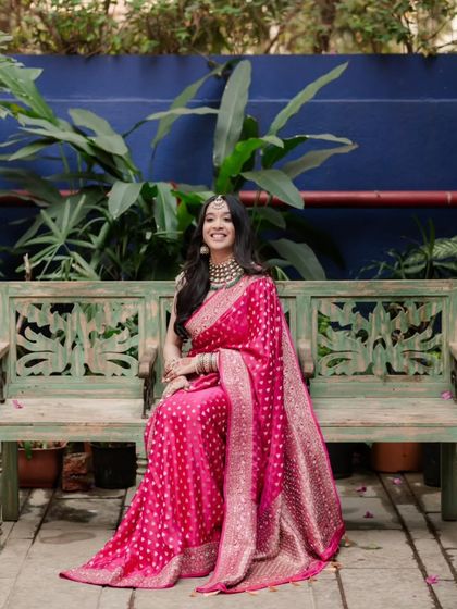 The bride, dressed in a vibrant pink saree, smiles brightly while seated on a rustic bench, a perfect blend of elegance and joy.