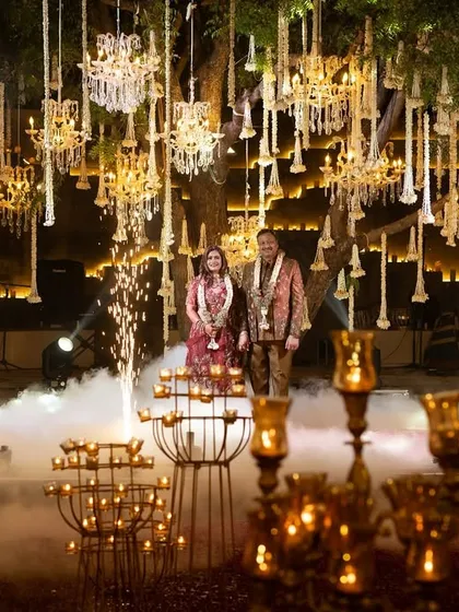 The couple stands on stage during their 25th-anniversary celebration, framed by cold pyrotechnics. The backdrop of hanging chandeliers and florals creates a truly spectacular and grand setting.