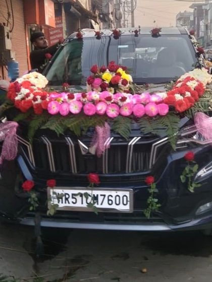 A frontal view of a decorated black Mahindra XUV 700. The thick floral garland across the front gives the car a grand and imposing presence.