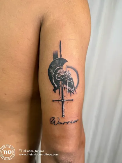 A powerful abstract tattoo representing the Spartan warrior spirit. The design combines a helmet, a sword, and the word "Warrior," symbolizing discipline, honor, and an unbreakable will.