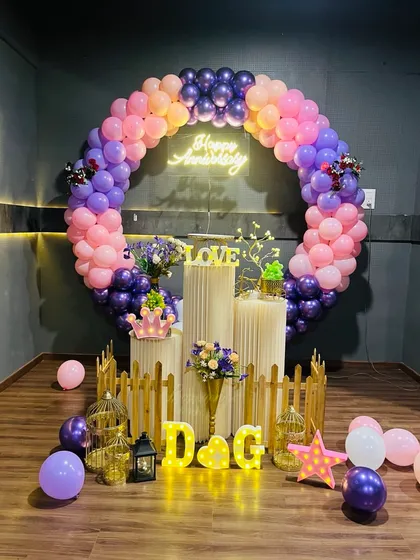 A beautiful anniversary decoration with a circular balloon arch in shades of pink and purple. The display is personalized with the couple's initials in marquee letters and includes a 'Happy Anniversary' neon sign.