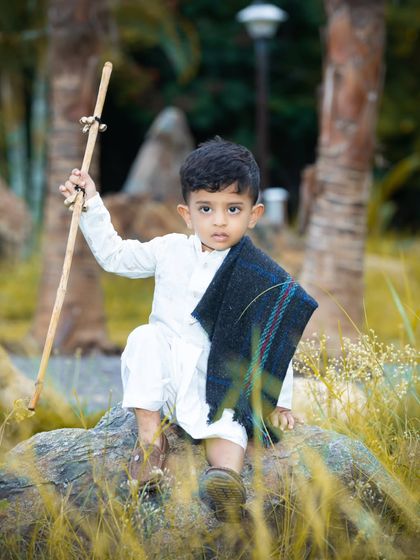 A young boy dressed in traditional white attire holds a staff, posing thoughtfully during an outdoor shoot in a park.