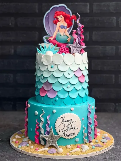 Let's plan an oceanic adventure with a custom mermaid cake. This two-tiered creation features beautiful teal and purple scales, an Ariel topper, and handcrafted sea elements, perfect for a magical under-the-sea birthday party.