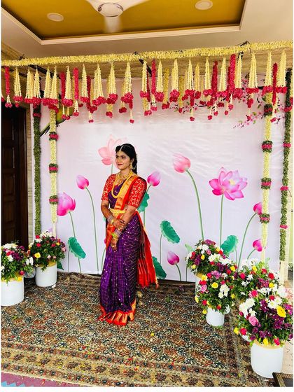 A full-length shot showcasing the entire look against the beautiful floral decor. The rich colors of her saree are enhanced by the classic makeup and simple, elegant hairstyle.