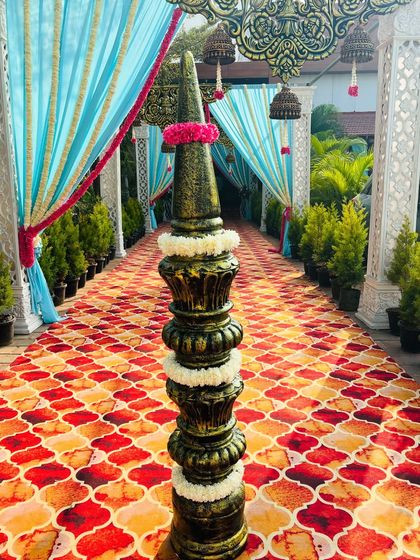 A traditional yet colorful walkway with light blue drapes, ornate pillars, and hanging temple bells. The patterned carpet and floral details add a touch of South Indian heritage to the entrance decor.