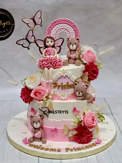 A lovely two-tier chocolate cake to welcome a princess. This design features soft pink tones, fondant teddy bears, a rainbow topper, and fresh floral accents for a sweet and elegant celebration.