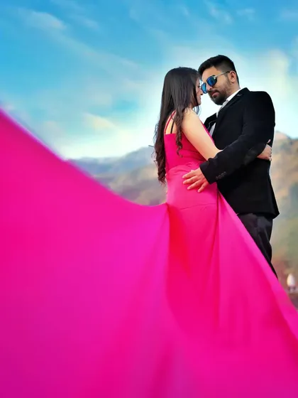 An intimate pre-wedding pose with the bride's long pink dress creating a dramatic foreground. The focus is on the couple's closeness against the vast mountain and sky backdrop.