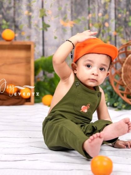 A fresh and fun orange-themed photoshoot for a baby boy. Dressed in cute overalls and a cap, he plays among scattered oranges in a rustic, light-filled studio setting.