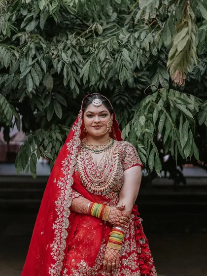 A bride in a heavy red lehenga, her colorful bangles a focal point, stands against the backdrop of our green gardens.