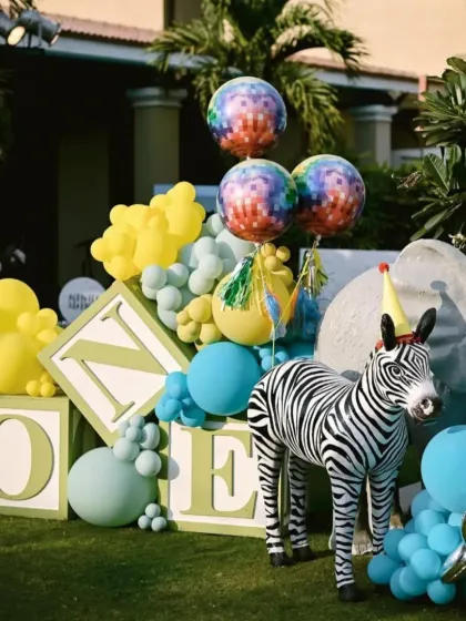 A fun fair theme with a twist. This setup for a first birthday includes large 'ONE' letter blocks and a friendly zebra prop amidst colorful balloons.