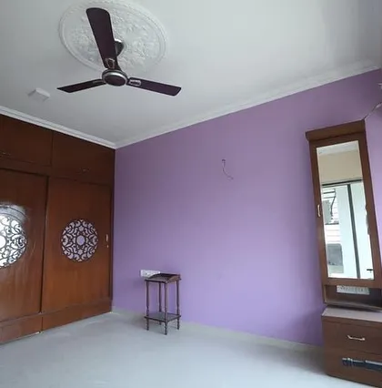 A spacious bedroom featuring a large wooden wardrobe with custom CNC-cut circular patterns on the doors. The room is painted in a calming lavender shade, complemented by a separate wooden dressing table.