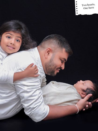 A playful and loving moment between a father and his two children. The older sister gives her dad a hug as he gazes at his newborn, showcasing the beautiful family dynamic.